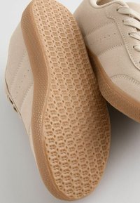 Beige ruskinds sneakers med runde snørebånd, tekstildetaljer og en gummisål med et honningcomb-mønster for greb og trækkraft.
