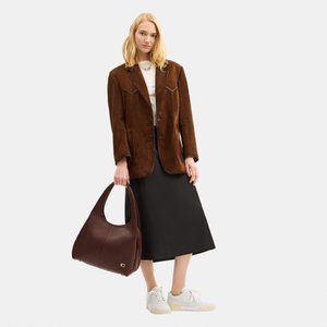 Vrouw draagt een bruine suède blazer, wit topje, zwarte midirok, witte sneakers, houdt een grote donkerbruine leren handtas vast.