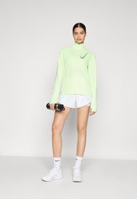 Light green long-sleeve athletic top with zipper, paired with white shorts. The model holds black dumbbells and wears white sneakers and socks.
