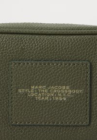 Olijfgroen leren crossbody tas met een gestructureerde afwerking. Voorzien van een gouden gestempeld label met merk, stijl, locatie en jaartal.