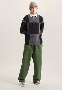 Chemise à manches longues bicolore noire et grise avec col, associée à un pantalon vert ample et des chaussures slip-on à carreaux. Bonnet tricoté porté.