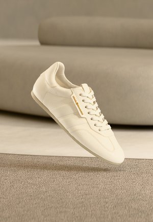 Svetlo béžová kožená teniska s textúrovaným povrchom, plochými šnúrkami a jemným logom, s elegantným dizajnom a minimalistickou siluetou.