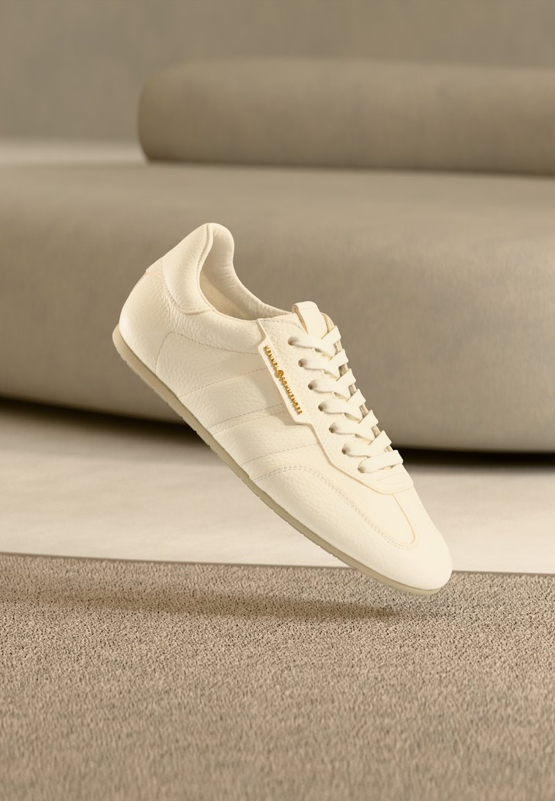 Ανοιχτό μπεζ δερμάτινο αθλητικό παπούτσι με υφή, επίπεδα κορδόνια και διακριτική σήμανση, με κομψό σχεδιασμό και μινιμαλιστική σιλουέτα.