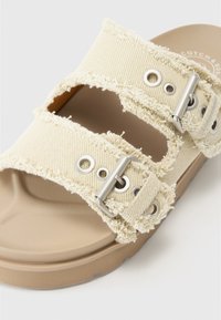 Beige kanka sandaalit, joissa on kaksi säädettävää hihnaa, revityt reunat ja metalliset yksityiskohdat. Teksturoitu pohja ja vaaleanruskea pohja.