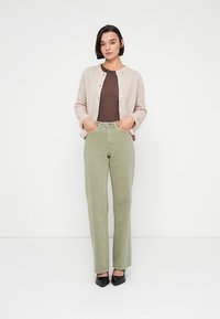 Γυναίκα που στέκεται με τα χέρια στις τσέπες, φορώντας μπεζ ζακέτα, καφέ μπλούζα, ανοιχτοπράσινο παντελόνι και μαύρα μυτερά παπούτσια σε λευκό φόντο.