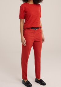 Röd texturerad kortärmad tröja med rundad halsringning, matchad med röda skräddarsydda byxor och svarta loafers. Svart bälte med silverdetalj.