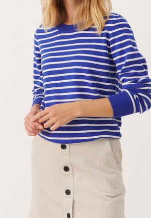 Kvinde iført en blå og hvidstribet langærmet trøje og beige kordnøj-skørt med knapper foran, stående mod en ensfarvet baggrund.