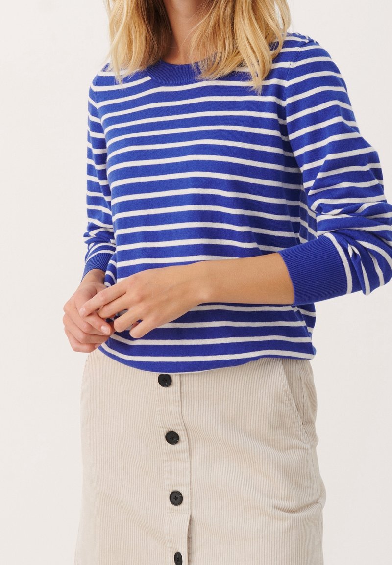 Femme portant un pull à manches longues à rayures bleues et blanches et une jupe en velours côtelé beige à boutons sur le devant, debout devant un fond uni.