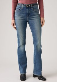 Γυναίκα που φοράει ξεθωριασμένο μπλε τζιν με ίσια μπάτσα και μαύρη ζώνη, ροζ ρόδου μακρυμάνικο τοπ και μαύρες μπότες με τακούνι.