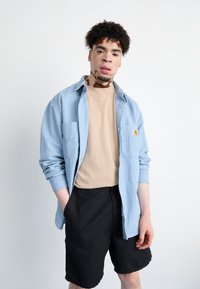 Hellblaues Hemd mit Knöpfen über einem beigen T-Shirt, kombiniert mit schwarzen Shorts. Das Hemd hat zwei vordere Taschen. Glatte Stofftexturen.