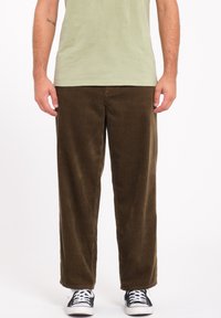 Pantalon en velours côtelé marron avec une coupe large et droite. Présente une texture à rayures verticales et une coupe décontractée. Associé à des baskets noires.