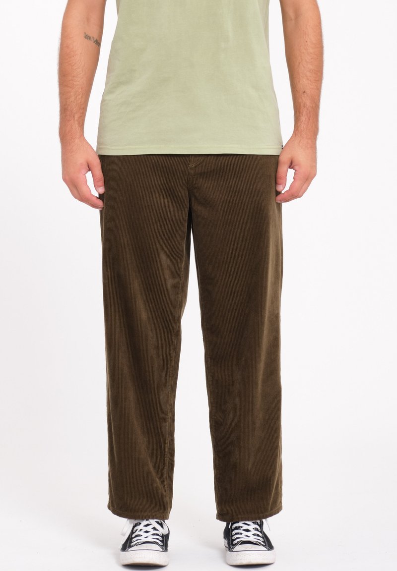 Pantalon en velours côtelé marron avec une coupe large et droite. Présente une texture à rayures verticales et une coupe décontractée. Associé à des baskets noires.