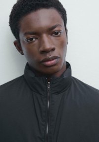 Jeune homme noir avec des cheveux bouclés courts portant une veste noire à zip, faisant face à l'avant avec une expression neutre sur un fond clair et uni.
