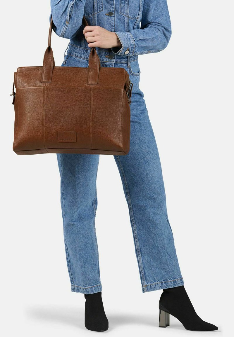 Persona in un completo di denim che tiene una grande borsa a tracolla in pelle marrone, indossando stivali neri con tacco alla caviglia, in piedi su uno sfondo bianco.