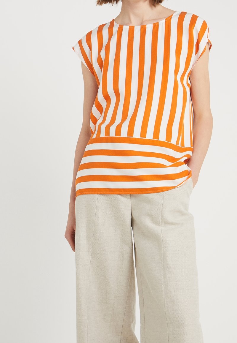 Femme porte un haut à manches courtes orange et blanc avec des rayures verticales et horizontales, associé à un pantalon large beige clair.