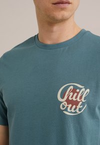 Zöld pamut póló, kerek nyakkivágással és elöl található grafikával, amin a "Chill out" szöveg szerepel fehér és piros betűkkel.