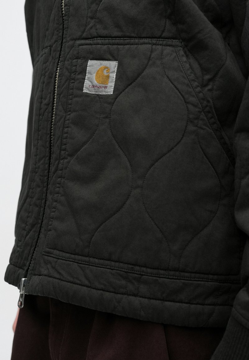 Black quilted jacket featuring a prominent logo patch, visible stitching patterns, and a zip closure at the hem.
