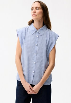 Kvinde med langt brunt hår iført en ærmeløs blå og hvid, vertikalt stribet skjorte med knapper, står med hænderne foldet og kigger til siden.