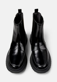 Botines negros con un acabado brillante y una correa decorativa en el frente, sobre un fondo gris claro.