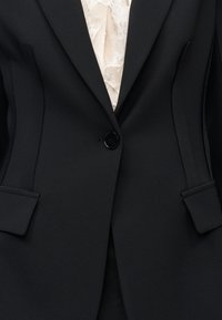 Μαύρο ραμμένο σακάκι με μονο κουμπί, από πάνω από κρεμ δαντελένιο μπλουζάκι με λεπτά ανθισμένα σχέδια και κοντό βολάν στη λαιμόκοψη.