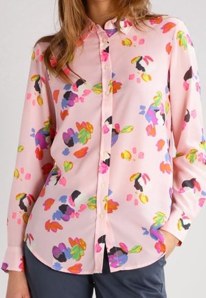 Jasnoróżowa, długorękawowa bluzka z kolorowym, abstrakcyjnym kwiatowym wzorem, z przodu na guziki, noszona z ciemnymi spodniami, dłoń w kieszeni.