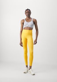 Lys grå sports-bh med tynne stropper, kombinert med lyse gule leggings i et glatt stoff, fullført med hvite treningssko.