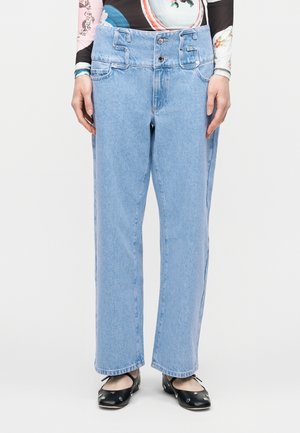 Ανοιχτό μπλε τζιν παντελόνι με φαρδιά γραμμή, διπλό κουμπί στη μέση και θηλιές για ζώνη, φορεμένο με μαύρες μπαλαρίνες και μακρυμάνικο τοπ με σχέδιο.