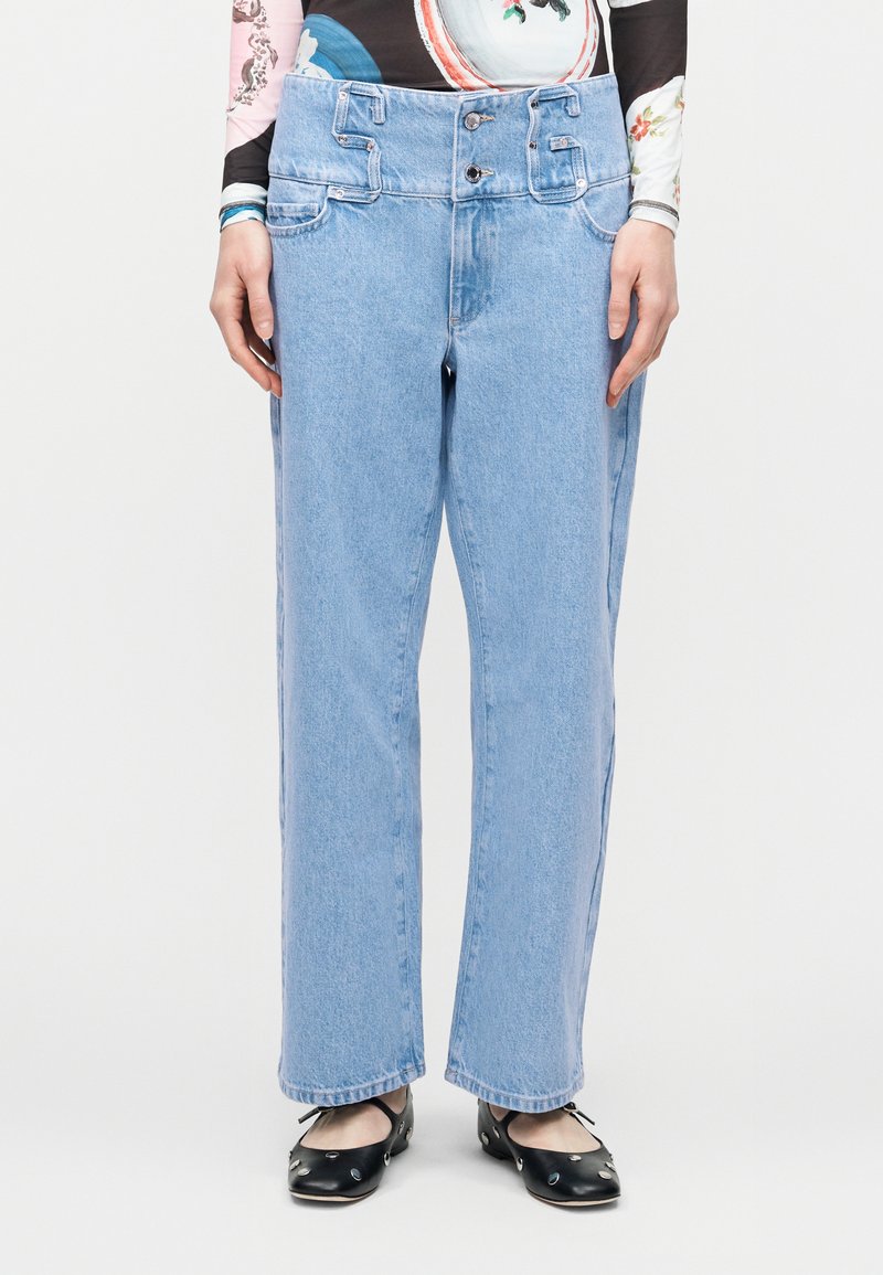 Ανοιχτό μπλε τζιν παντελόνι με φαρδιά γραμμή, διπλό κουμπί στη μέση και θηλιές για ζώνη, φορεμένο με μαύρες μπαλαρίνες και μακρυμάνικο τοπ με σχέδιο.