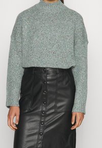 Suéter de punto verde claro con motas multicolores, cuello alto y corte holgado, combinado con falda de cuero negra con detalles de botones.