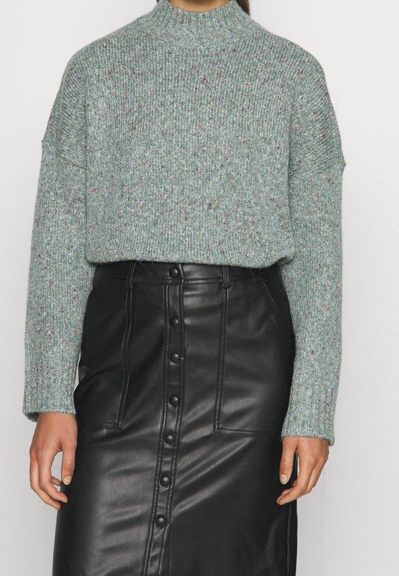 Suéter de punto verde claro con motas multicolores, cuello alto y corte holgado, combinado con falda de cuero negra con detalles de botones.