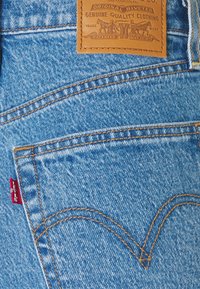 Jeans de mezclilla azul con un parche de cuero que presenta texto en relieve, costuras naranjas en contraste y una etiqueta de marca roja visible en la costura del bolsillo.