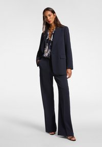 Traje azul marino con una blazer ajustada y pantalones de pierna ancha, superpuesto a una blusa estampada. Presenta una tela suave y un diseño minimalista.