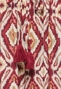 Tissu à motif géométrique rouge, orange et beige avec deux pompons rouges suspendus au centre.