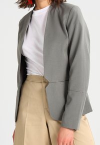 Šedý blazer s čistým, strukturovaným designem, s dlouhými rukávy, šálovým límcem a asymetrickým předním lemem. Nositý přes bílou košili.