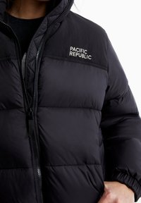 Doudoune noire avec panneaux cousus, col montant et fermeture éclair. Logo "PACIFIC REPUBLIC" en blanc sur le haut de l'avant.