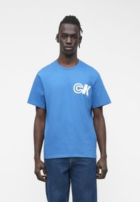 Μπλε βαμβακερό μπλουζάκι με στρογγυλή λαιμόκοψη και κοντά μανίκια, με λευκό λογότυπο "CK" στο αριστερό στήθος. Συνδυασμένο με τζιν τζιν.