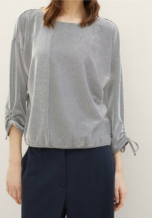 Femme portant un chemisier à rayures noires et blanches à manches longues froncées et un pantalon bleu marine, debout devant un fond uni.