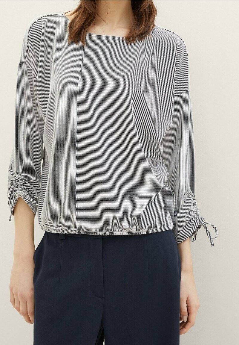 Kobieta ubrana w czarno-białą bluzkę w paski z marszczonymi rękawami i granatowe spodnie, stojąca na jednolitym tle.