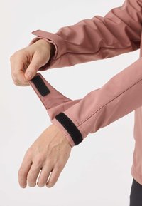 Rankos reguliuoja užsegamą lipnią juostelę prie švelniai rožinės ilgarankovės striukės rankogalių, fone matosi šviesi ir paprasta aplinka.