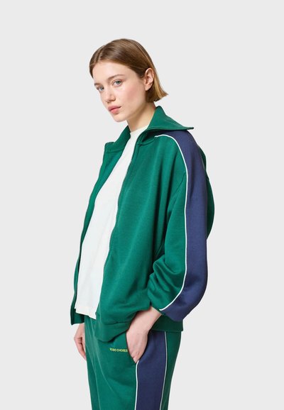 Zelena i tamnoplava majica s zipom i bijelim obrubom, s visokim ovratnikom i mekanim materijalom. U paru s odgovarajućim trenirkama.