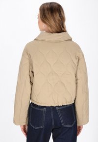 Beige cropped quiltet jakke med bred krave og elastisk talje. Bagsiden har en glat tekstur med et subtilt bølget mønster.