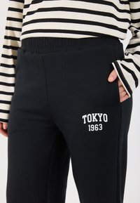 Czarne dresy z elastycznym paskiem w talii, z białym nadrukiem "TOKYO 1963" z boku. Materiał wydaje się miękki i gładki.