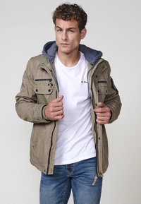 Khaki bunda so šedou kapucňou, so viacerými zipsovými a gombíkovými vreckami, textúrovanou látkou a zapínaním na zips, oblečená na bielom tričku.