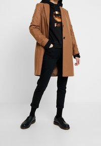 Persona vistiendo un abrigo color camello sobre una sudadera negra con logo de color cobre, pantalones negros arremangados, calcetines negros y zapatos negros de cordones sobre un fondo plano.