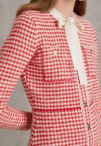Czerwona i kremowa marynarka w kratę houndstooth z długimi rękawami, dwoma kieszeniami na piersiach oraz białą kokardą przy kołnierzu. Dodatki z metalowych guzików.