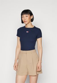 Mörkblå figursydd t-shirt med korta ärmar och rund halsringning, med en liten logotyp i röd och vit. Tillsammans med beige veckade shorts.