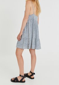 Blaues Blümchenmuster-Kleid mit Spaghettiträgern, gerafftem Bund und weit ausgestelltem Saum. Kombiniert mit schwarzen Sandalen mit Riemen und verstellbaren Schnallen.