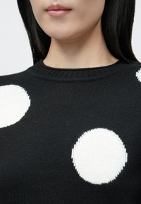 Jersey negro con cuello redondo, con grandes lunares blancos en textura de punto. Vista en primer plano del diseño y detalles de la tela.