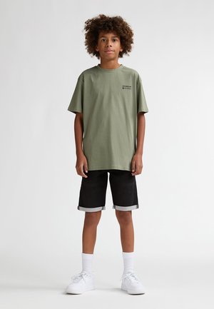 Tricou verde măsliniu din bumbac, cu mâneci scurte, combinat cu pantaloni scurți negri din denim cu tivuri rulate și adidași albi cu șosete albe.