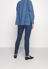 Camisa de mezclilla de corte relajado, color azul, con puños abotonados, combinada con jeans ajustados de color azul oscuro y zapatos negros sin cordones.